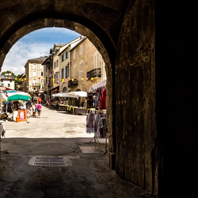 Passage voûté menant à la rue commerçante de Mont-Louis animée, avec ses étals, ses promeneurs et ses façades en pierre typiques d’une cité fortifiée.