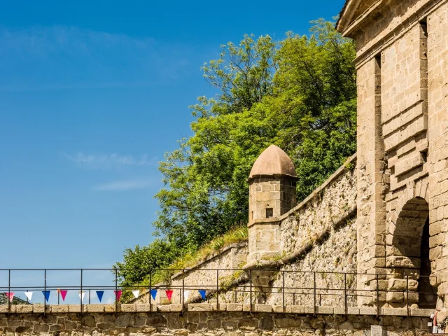 Entrée fortifiée de Mont-Louis avec sa guérite de pierre et son pont décoré de fanions colorés sous un ciel bleu d’été.