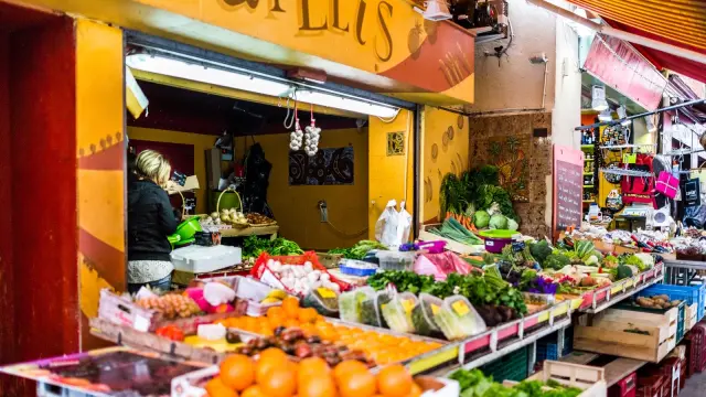 Étal coloré d’un marché couvert avec fruits, légumes et produits frais, tenu par une commerçante.