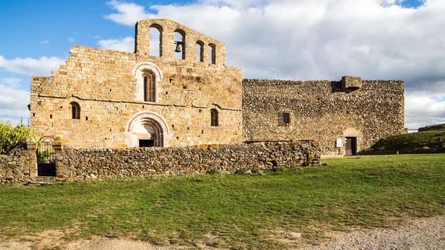 Église romane en pierre dorée avec clocher à arcades et bâtiment fortifié attenant, sous un ciel bleu parsemé de nuages.