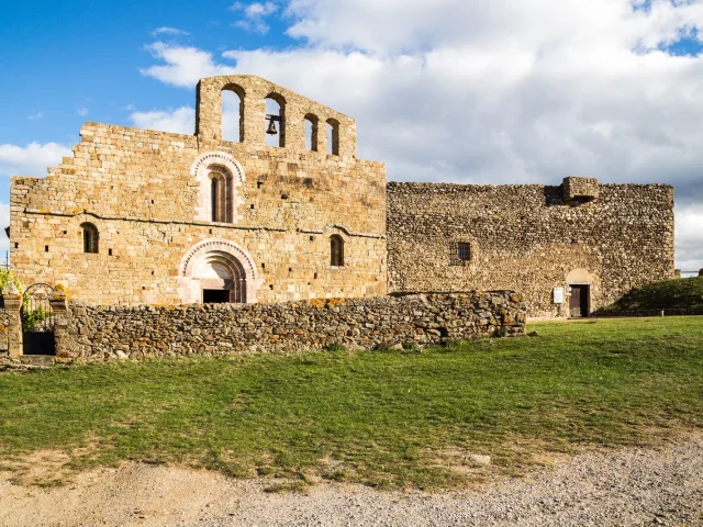 Église romane en pierre dorée avec clocher à arcades et bâtiment fortifié attenant, sous un ciel bleu parsemé de nuages.