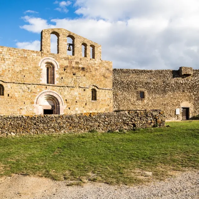 Église romane en pierre dorée avec clocher à arcades et bâtiment fortifié attenant, sous un ciel bleu parsemé de nuages.