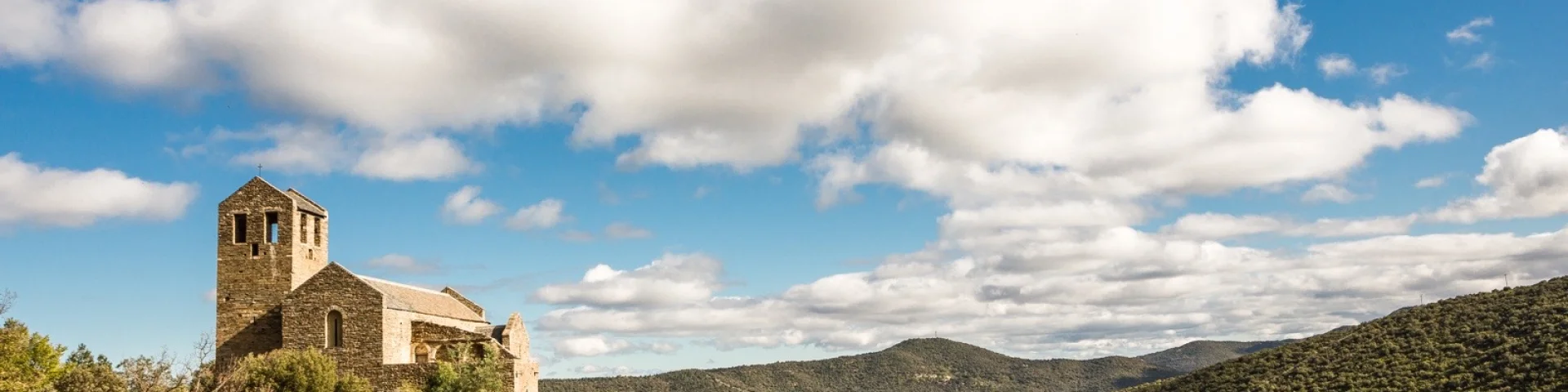 Église romane en pierre perchée sur une colline verdoyante, entourée de garrigue et de montagnes sous un ciel bleu parsemé de nuages blancs.