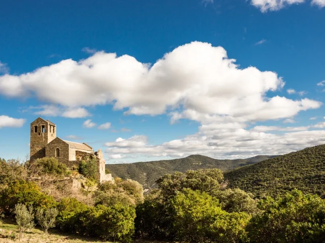 Église romane en pierre perchée sur une colline verdoyante, entourée de garrigue et de montagnes sous un ciel bleu parsemé de nuages blancs.