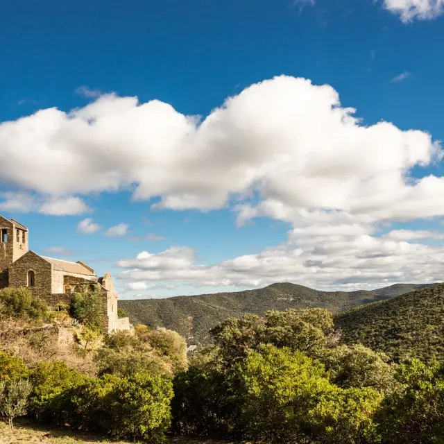 Église romane en pierre perchée sur une colline verdoyante, entourée de garrigue et de montagnes sous un ciel bleu parsemé de nuages blancs.
