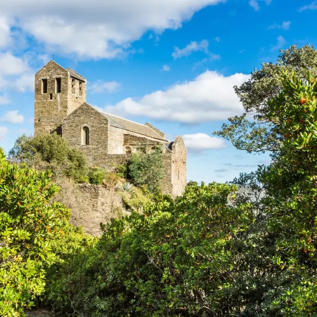 Ancienne abbaye romane en pierre perchée sur une colline verdoyante, entourée de végétation méditerranéenne sous un ciel bleu.