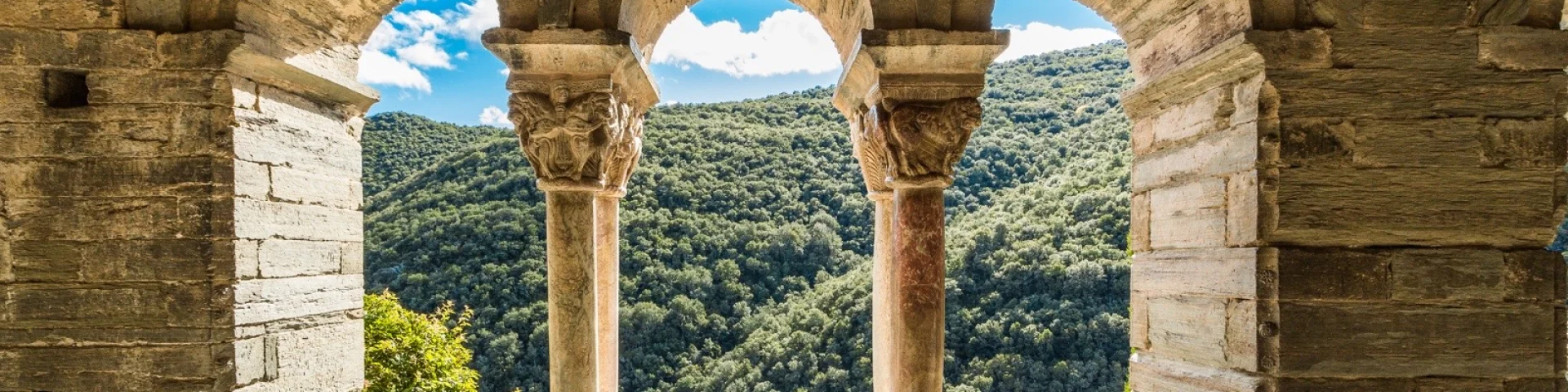 Vue à travers les arcades d’un cloître roman en pierre, ouvrant sur les collines verdoyantes d’une vallée ensoleillée.