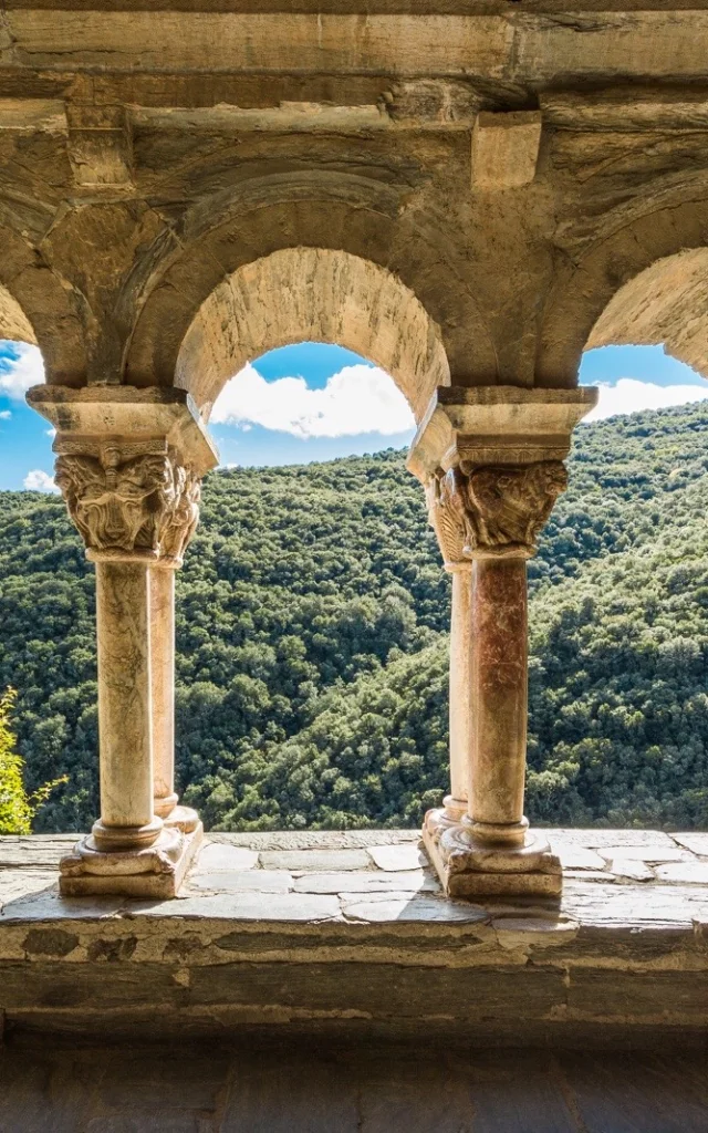 Vue à travers les arcades d’un cloître roman en pierre, ouvrant sur les collines verdoyantes d’une vallée ensoleillée.