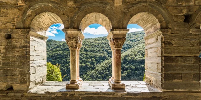 Vue à travers les arcades d’un cloître roman en pierre, ouvrant sur les collines verdoyantes d’une vallée ensoleillée.
