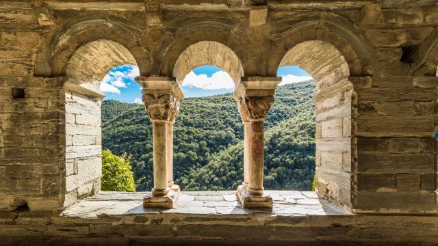 Vue à travers les arcades d’un cloître roman en pierre, ouvrant sur les collines verdoyantes d’une vallée ensoleillée.