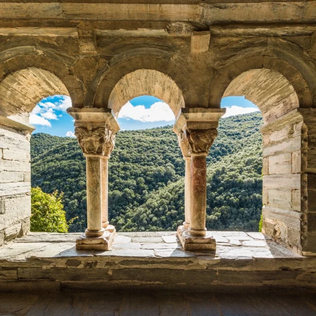 Vue à travers les arcades d’un cloître roman en pierre, ouvrant sur les collines verdoyantes d’une vallée ensoleillée.