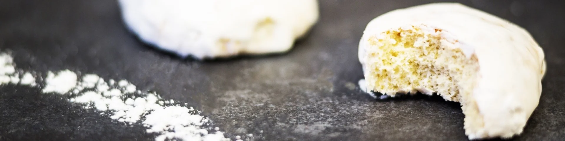 Deux rousquilles catalanes couvertes de glaçage blanc, posées sur une surface sombre saupoudrée de sucre glace, dont l’une est entamée.