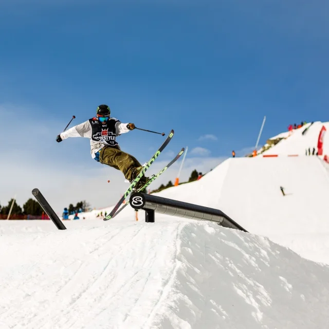 Skieur freestyle effectuant une figure aérienne sur un rail dans le snowpark enneigé de Font-Romeu, sous un ciel bleu.
