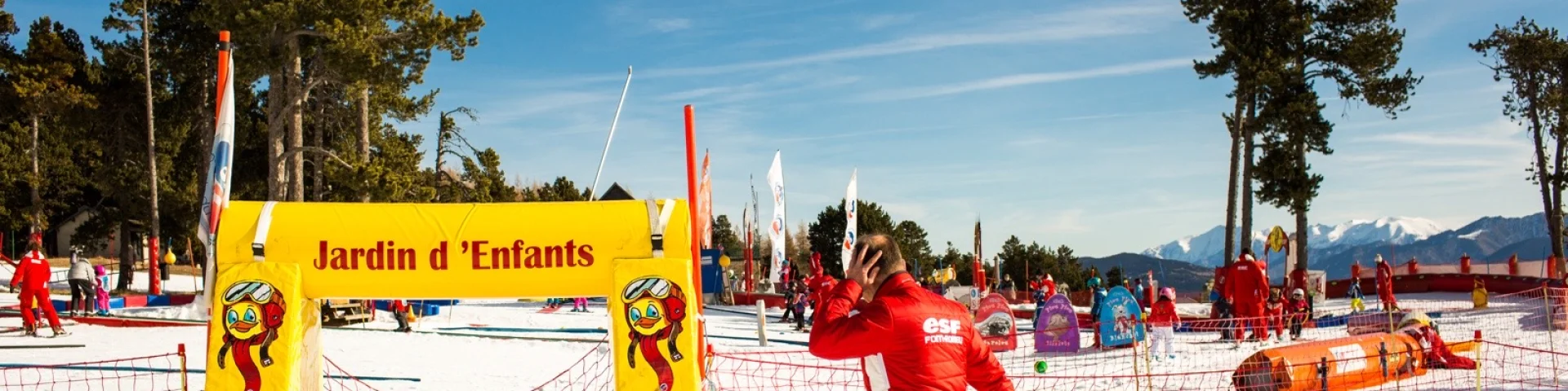 Moniteur de ski en combinaison rouge passant devant l’entrée du Jardin d’Enfants de l’ESF à Font-Romeu, sous un ciel bleu et face aux montagnes enneigées.
