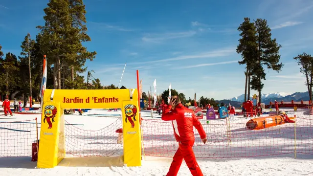 Moniteur de ski en combinaison rouge passant devant l’entrée du Jardin d’Enfants de l’ESF à Font-Romeu, sous un ciel bleu et face aux montagnes enneigées.