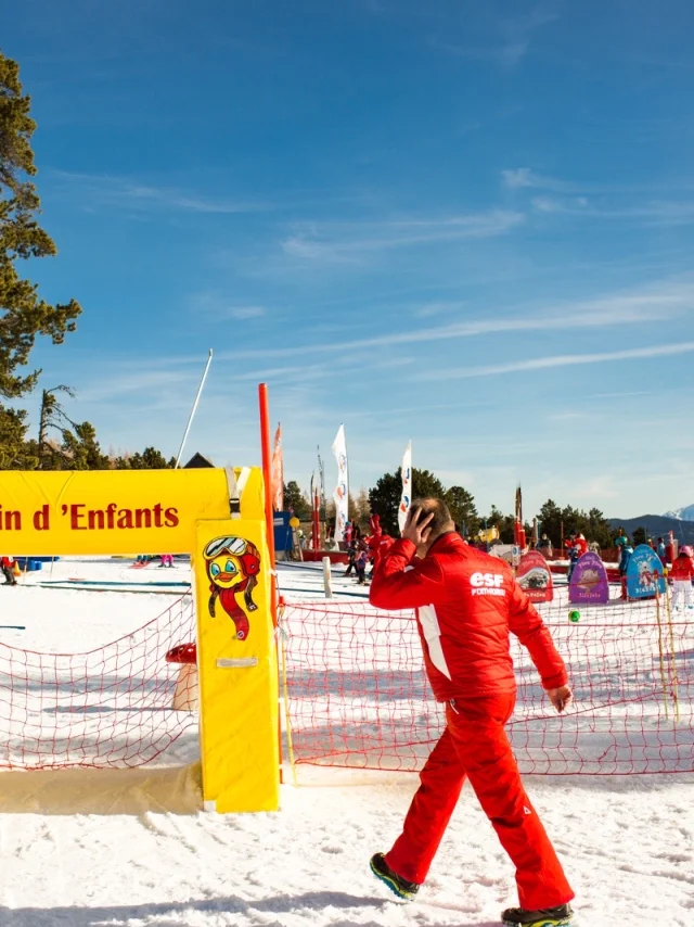 Moniteur de ski en combinaison rouge passant devant l’entrée du Jardin d’Enfants de l’ESF à Font-Romeu, sous un ciel bleu et face aux montagnes enneigées.