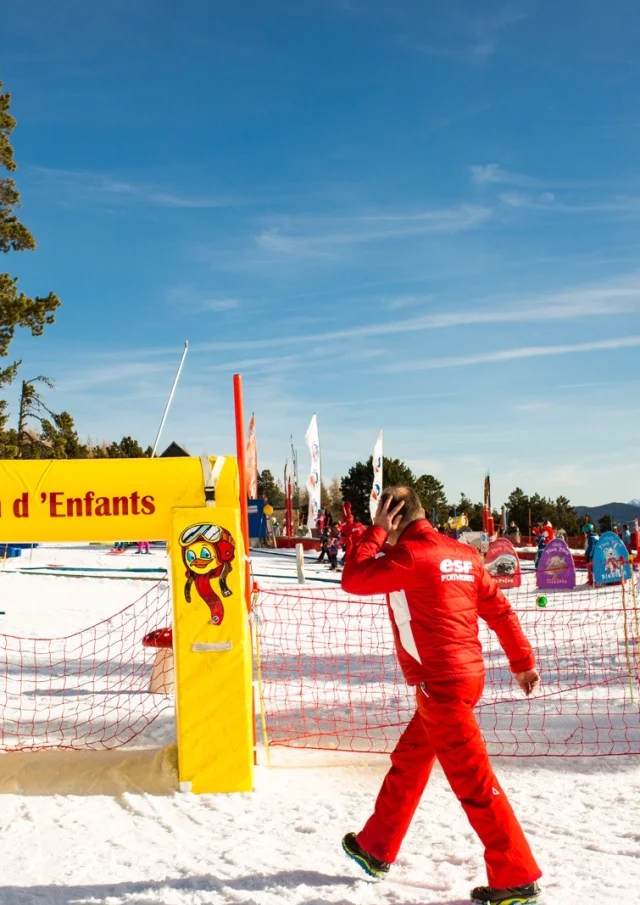Moniteur de ski en combinaison rouge passant devant l’entrée du Jardin d’Enfants de l’ESF à Font-Romeu, sous un ciel bleu et face aux montagnes enneigées.