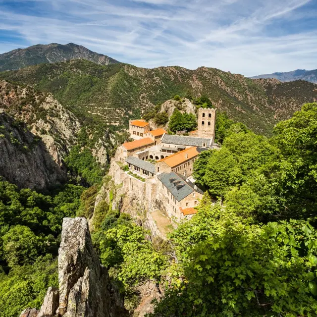 Le monastère de Saint-Martin-du-Canigó perché sur un piton rocheux, entouré de forêts verdoyantes et des montagnes du massif du Canigó sous un ciel bleu.