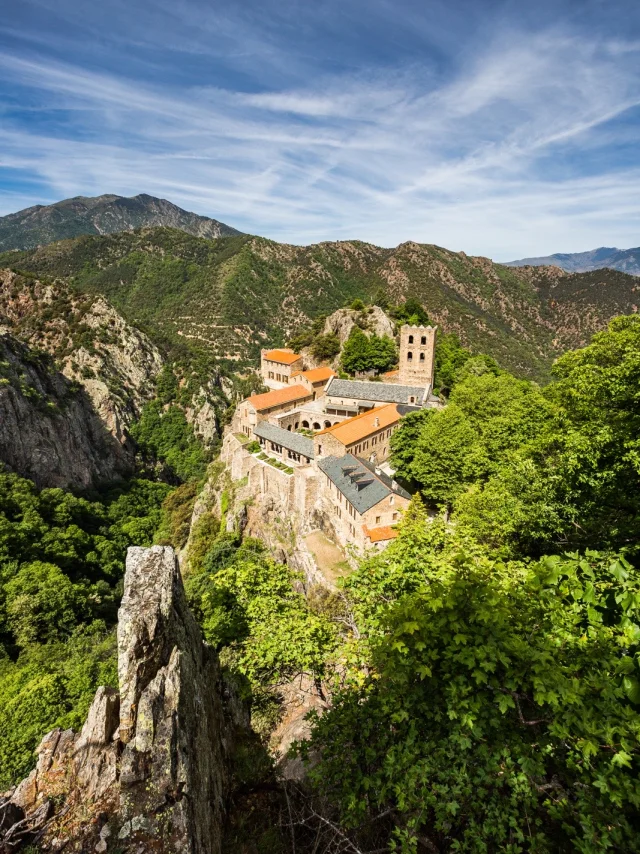 Le monastère de Saint-Martin-du-Canigó perché sur un piton rocheux, entouré de forêts verdoyantes et des montagnes du massif du Canigó sous un ciel bleu.