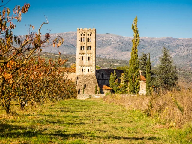 Abbaye romane Sainte-Marie d’Arles-sur-Tech entourée de vergers et de collines, sous un ciel bleu limpide.