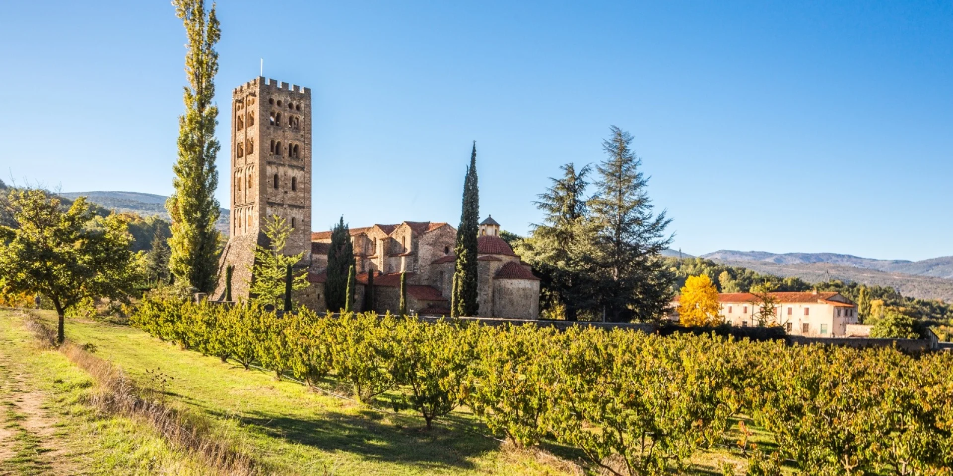 Vue de l’abbaye Saint-Michel de Cuxa entourée de vergers, au pied des montagnes, sous un ciel bleu limpide.