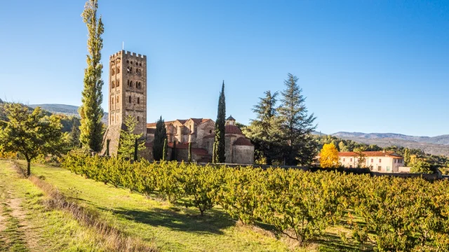 Vue de l’abbaye Saint-Michel de Cuxa entourée de vergers, au pied des montagnes, sous un ciel bleu limpide.