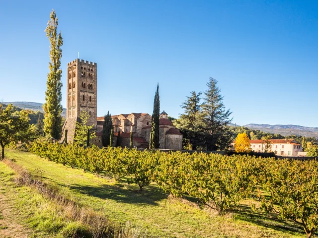 Vue de l’abbaye Saint-Michel de Cuxa entourée de vergers, au pied des montagnes, sous un ciel bleu limpide.