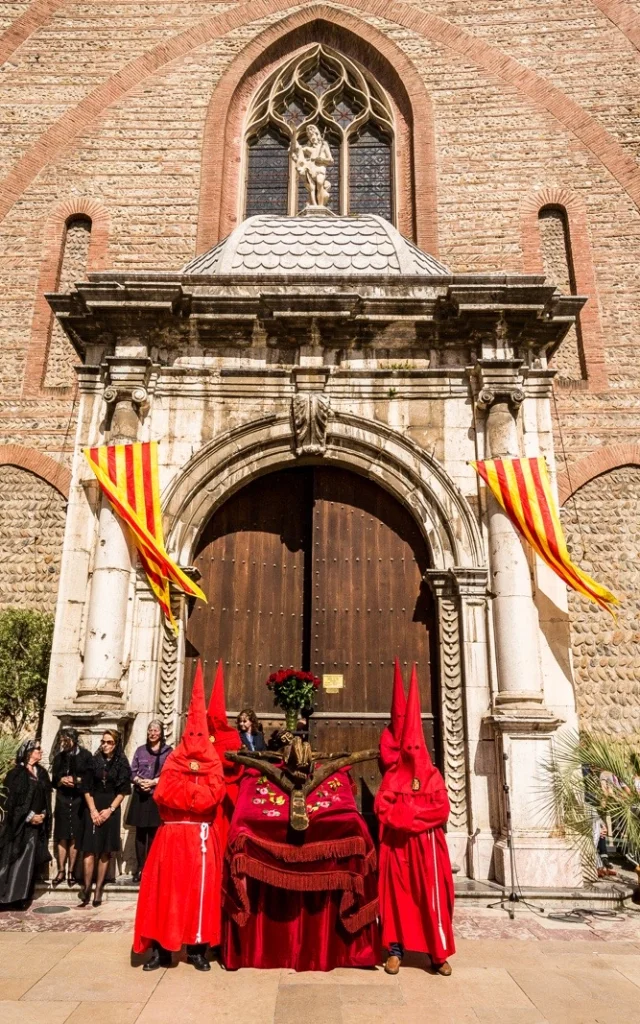 Devant l’église Saint-Jacques de Perpignan, des pénitents vêtus de rouge participent à la Procession de la Sanch, sous les drapeaux catalans et devant un public rassemblé.