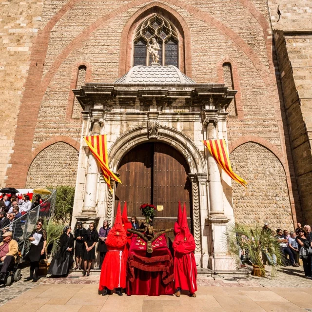Devant l’église Saint-Jacques de Perpignan, des pénitents vêtus de rouge participent à la Procession de la Sanch, sous les drapeaux catalans et devant un public rassemblé.