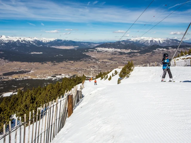 Skieur utilisant un téléski sur une piste enneigée, avec vue panoramique sur la vallée de Cerdagne et les montagnes des Pyrénées-Orientales sous un ciel bleu.