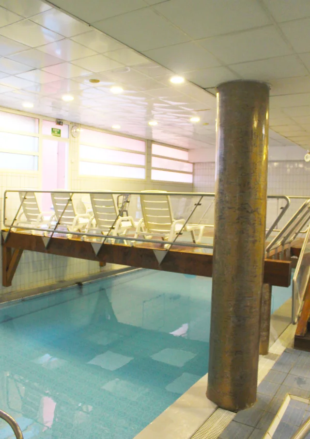 Piscine intérieure d’un centre thermal avec pont en bois et chaises longues pour la détente.