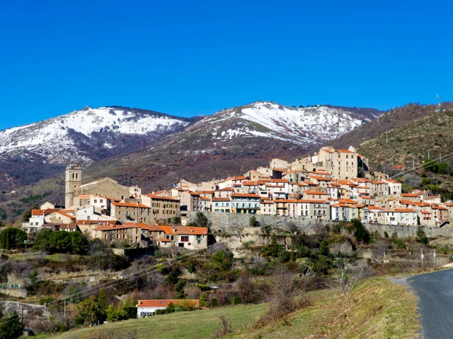 Vue du village perché de Vernet-les-Bains, aux maisons colorées et toits de tuiles rouges, dominé par les montagnes enneigées sous un ciel bleu éclatant.