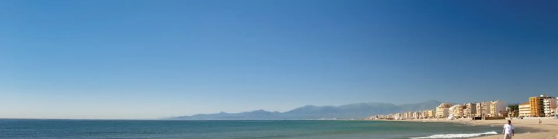 Plage de sable fin au Barcarès, bordée par la mer Méditerranée et les immeubles du front de mer, avec les montagnes du Canigó à l’horizon.