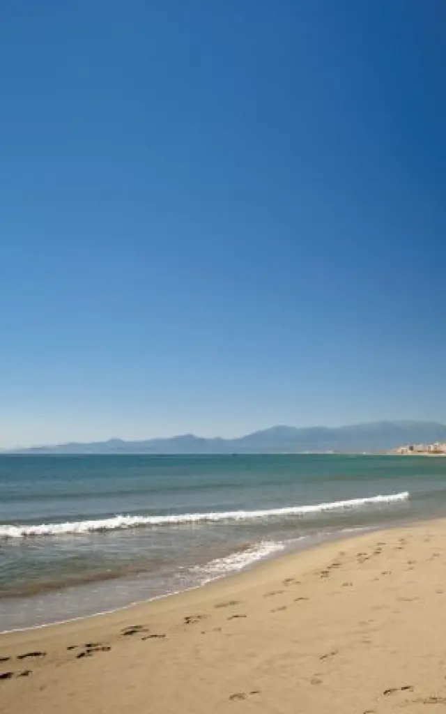 Plage de sable fin au Barcarès, bordée par la mer Méditerranée et les immeubles du front de mer, avec les montagnes du Canigó à l’horizon.
