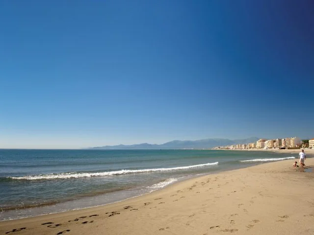 Plage de sable fin au Barcarès, bordée par la mer Méditerranée et les immeubles du front de mer, avec les montagnes du Canigó à l’horizon.