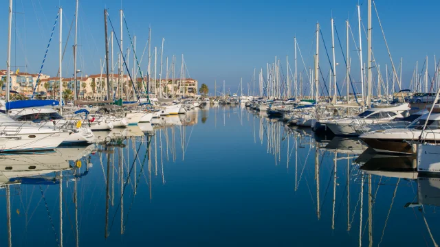 Alignement de voiliers et de bateaux de plaisance amarrés dans un port calme, leurs mats se reflétant dans l’eau sous un ciel bleu.