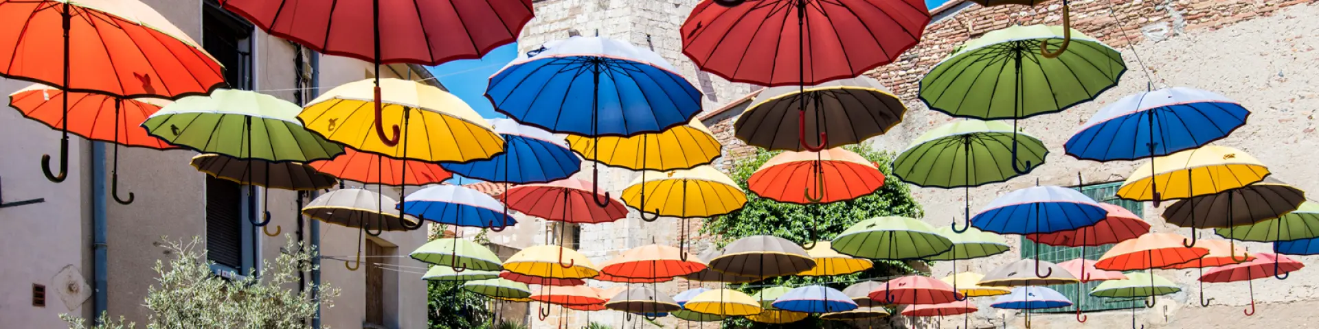 Installation artistique composée de nombreux parapluies colorés suspendus au-dessus d’une placette ensoleillée bordée de murs en pierre et de plantes méditerranéennes.