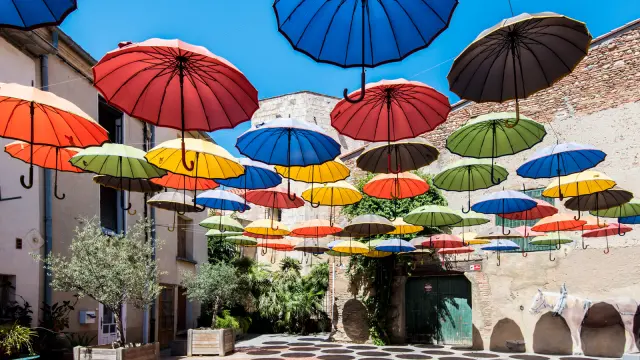 Installation artistique composée de nombreux parapluies colorés suspendus au-dessus d’une placette ensoleillée bordée de murs en pierre et de plantes méditerranéennes.