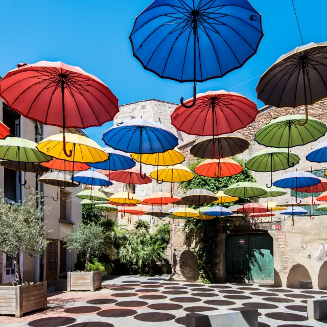 Installation artistique composée de nombreux parapluies colorés suspendus au-dessus d’une placette ensoleillée bordée de murs en pierre et de plantes méditerranéennes.
