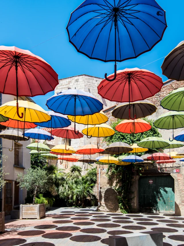 Installation artistique composée de nombreux parapluies colorés suspendus au-dessus d’une placette ensoleillée bordée de murs en pierre et de plantes méditerranéennes.