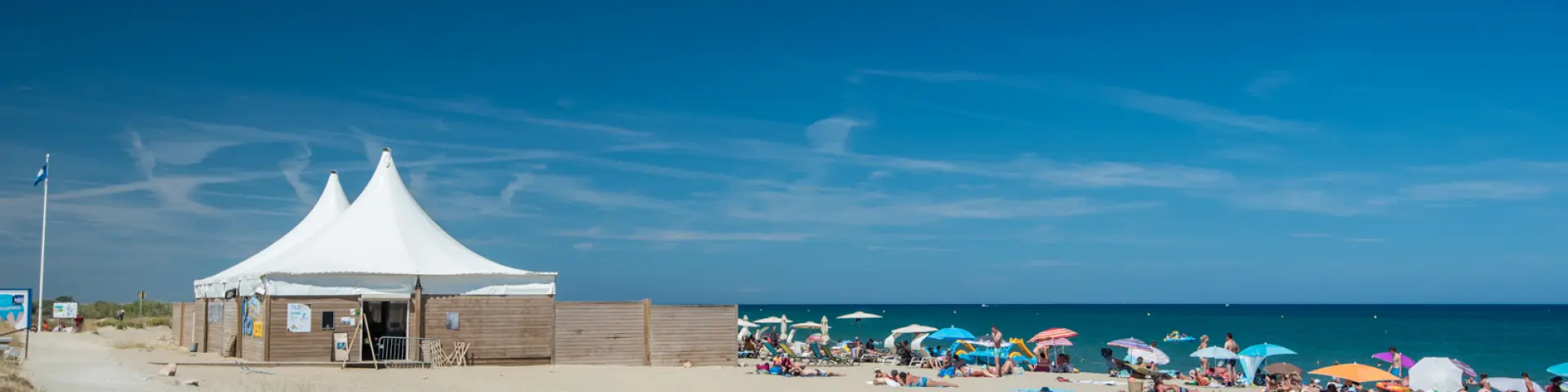 Plage de sable fin bordée par la mer Méditerranée, avec des vacanciers sous des parasols colorés et une grande tente blanche installée sur le sable.