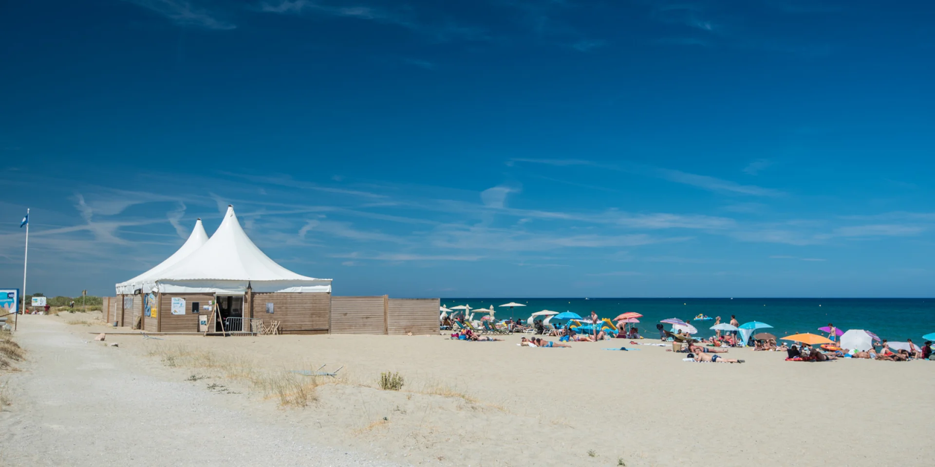 Plage de sable fin bordée par la mer Méditerranée, avec des vacanciers sous des parasols colorés et une grande tente blanche installée sur le sable.