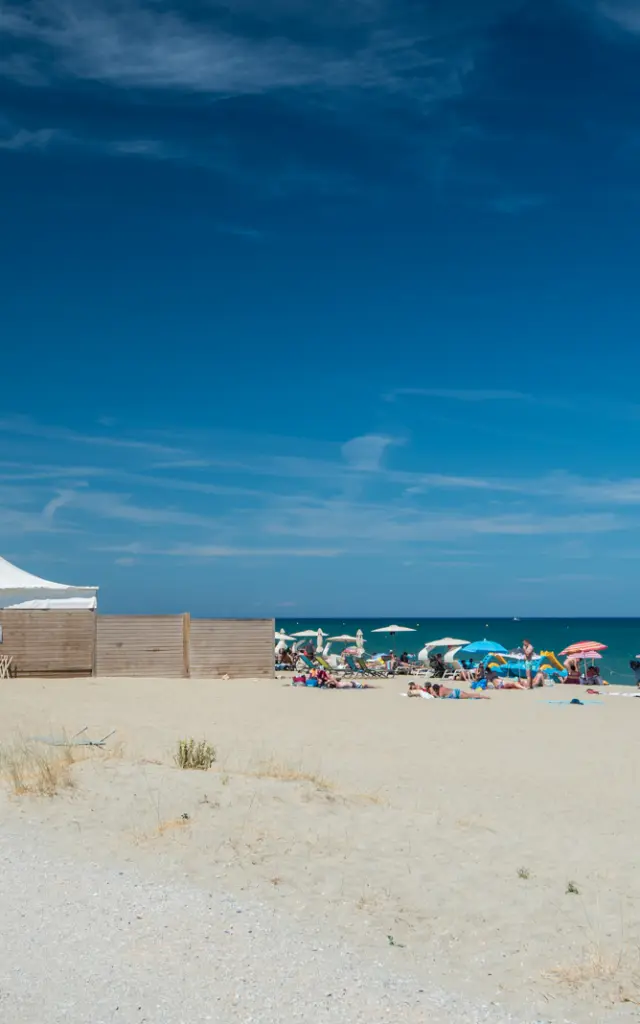 Plage de sable fin bordée par la mer Méditerranée, avec des vacanciers sous des parasols colorés et une grande tente blanche installée sur le sable.