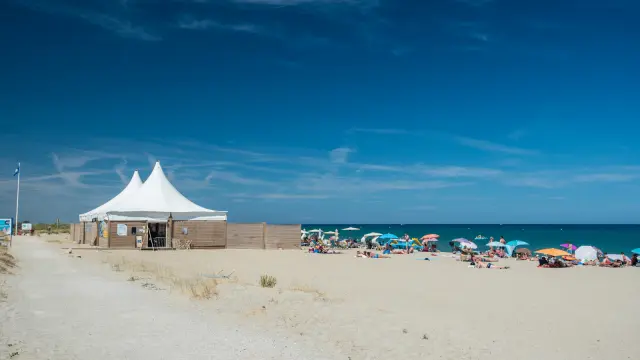 Plage de sable fin bordée par la mer Méditerranée, avec des vacanciers sous des parasols colorés et une grande tente blanche installée sur le sable.