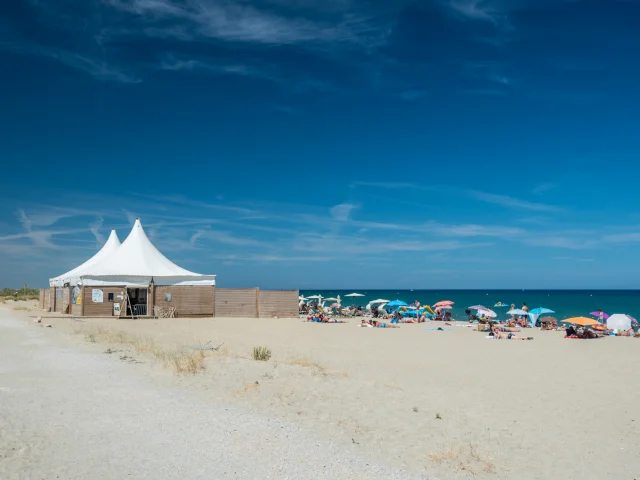 Plage de sable fin bordée par la mer Méditerranée, avec des vacanciers sous des parasols colorés et une grande tente blanche installée sur le sable.