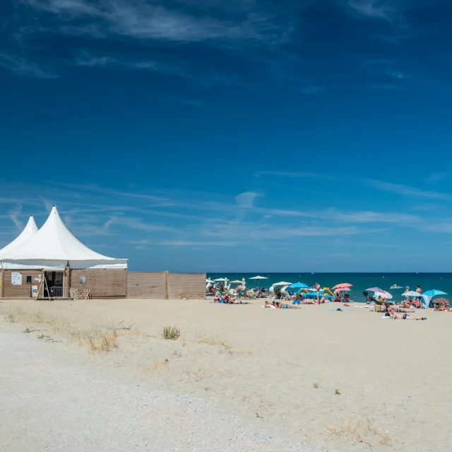 Plage de sable fin bordée par la mer Méditerranée, avec des vacanciers sous des parasols colorés et une grande tente blanche installée sur le sable.