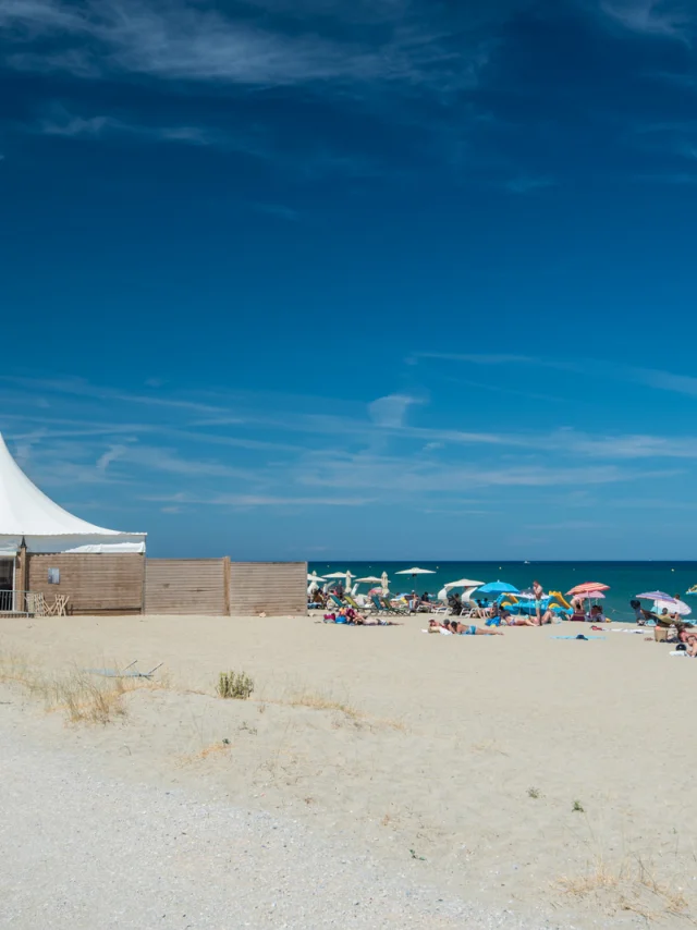 Plage de sable fin bordée par la mer Méditerranée, avec des vacanciers sous des parasols colorés et une grande tente blanche installée sur le sable.