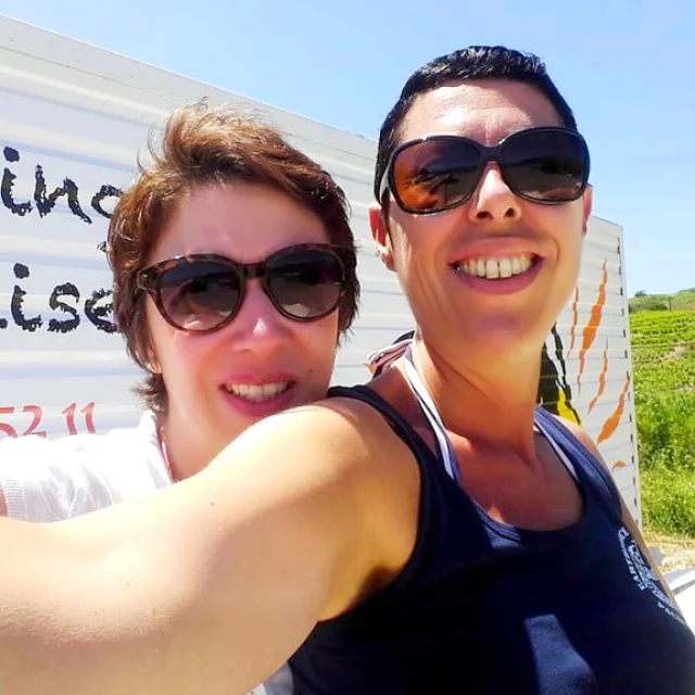 Deux femmes souriantes portant des lunettes de soleil prennent un selfie en extérieur, devant une enseigne indiquant une base de paddle.