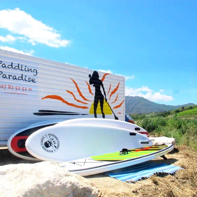 Planche de stand up paddle posée au sol devant une remorque signalant « Paddling Paradise », sur fond de vignes et de montagnes sous un ciel bleu.
