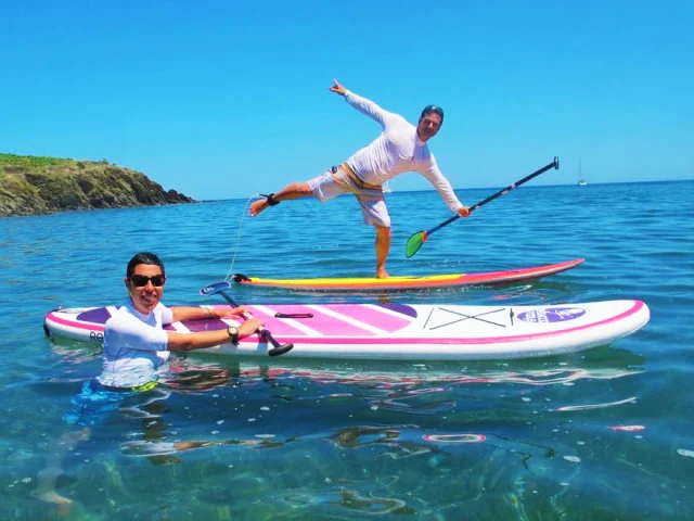 Deux personnes s’amusent sur leurs planches de stand up paddle dans une mer turquoise, sous un ciel bleu éclatant, au large des criques rocheuses de la Côte Vermeille.